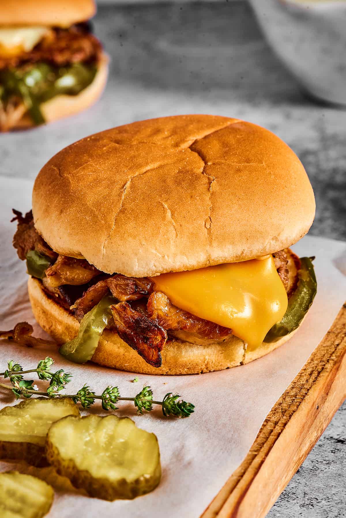 A steak sandwich on a hamburger bun with dill pickle chips and fresh thyme.