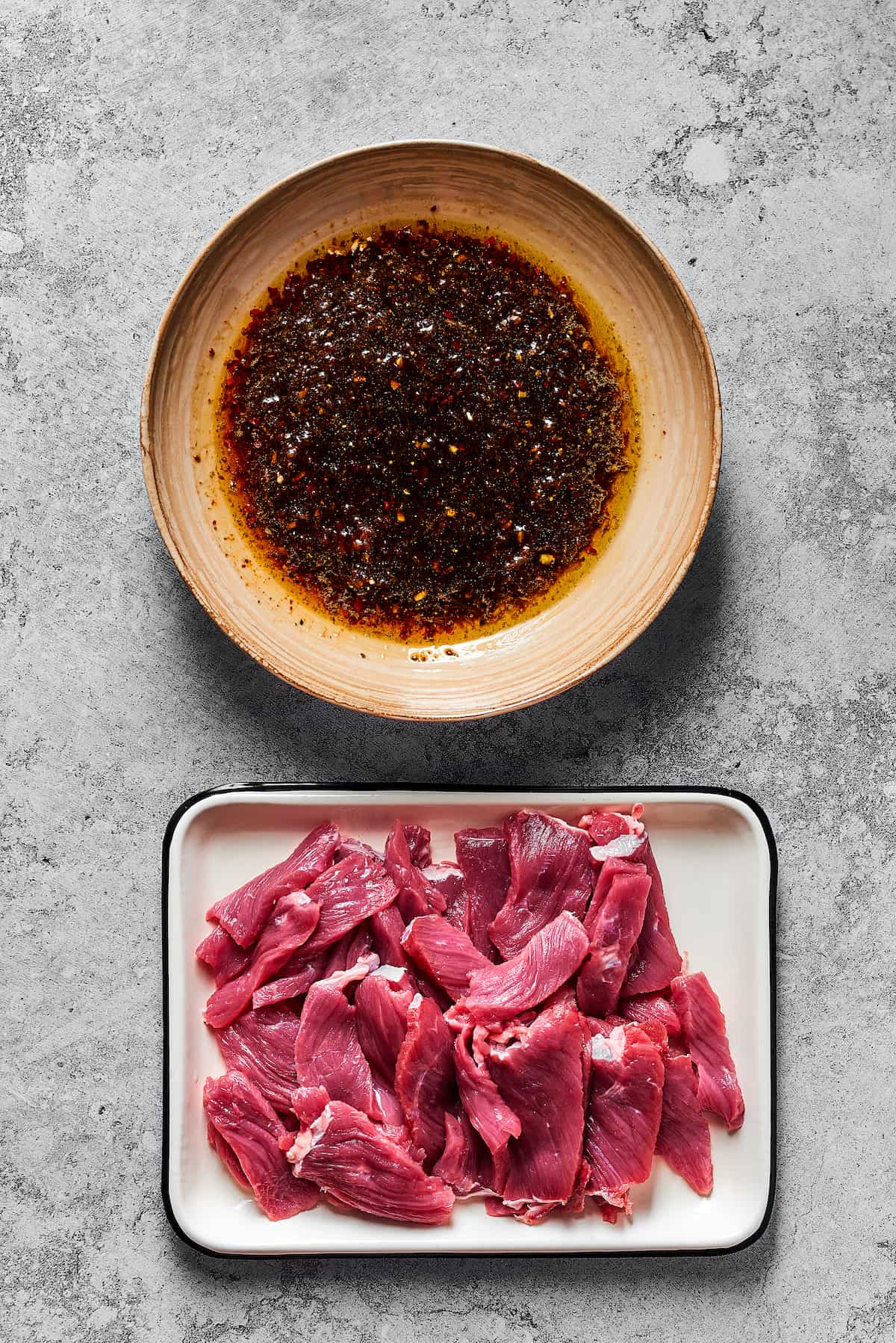 A bowl of marinade next to an enamel tray of sliced steak.