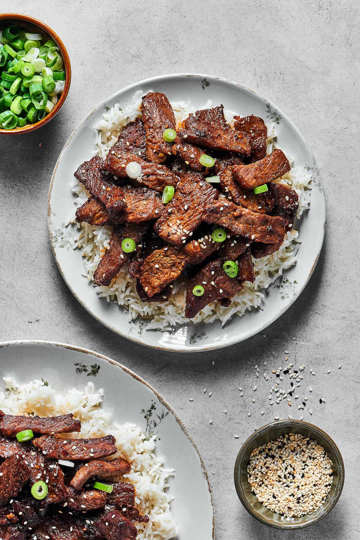 Cooked steak slices on a bed of rice, with garnishes of green onion and sesame seeds.