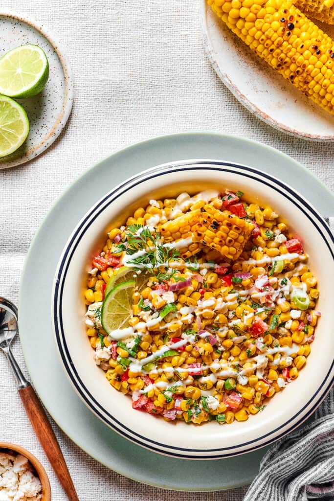 Mexican street corn asalad in a serving bowl, garnished with dressing and lime wedges.