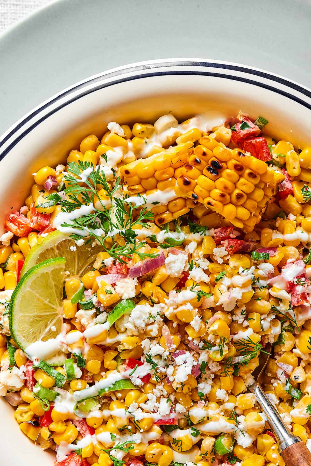 Close-up shot of grilled corn salad with creamy dressing, lime wedges, and chopped herbs.