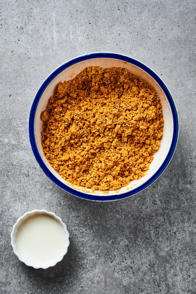 A crumbly mixture of graham crackers, butter, and sugar in a bowl. A small dish of milk is near the bowl.