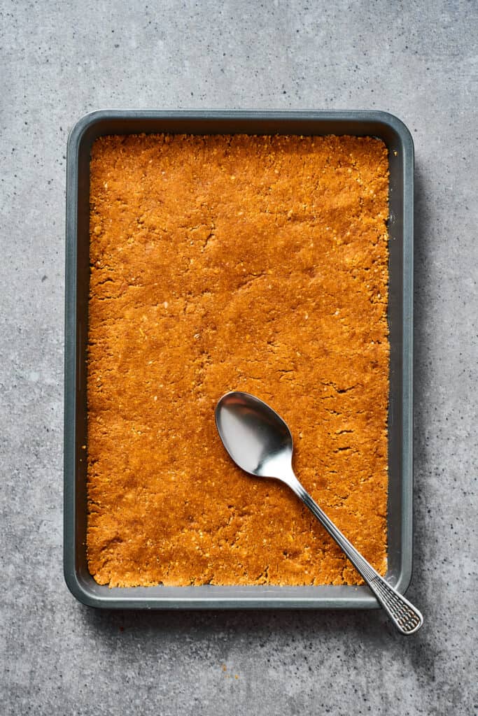 A cookie crust pressed into a baking dish. A spoon is resting on the crust.