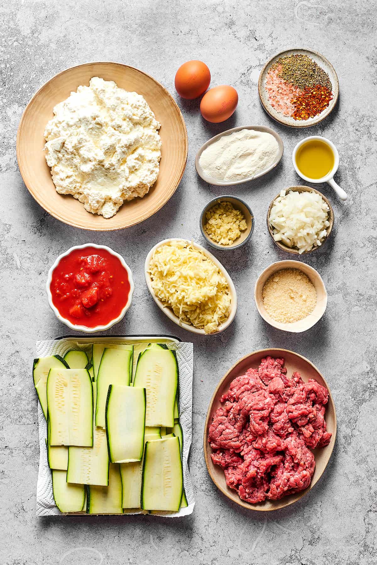 From top left: Cottage cheese, eggs, seasonings, four, olive oil, garlic, diced onion, crushed tomatoes, mozzarella, Parmesan, zucchini slices, ground beef.
