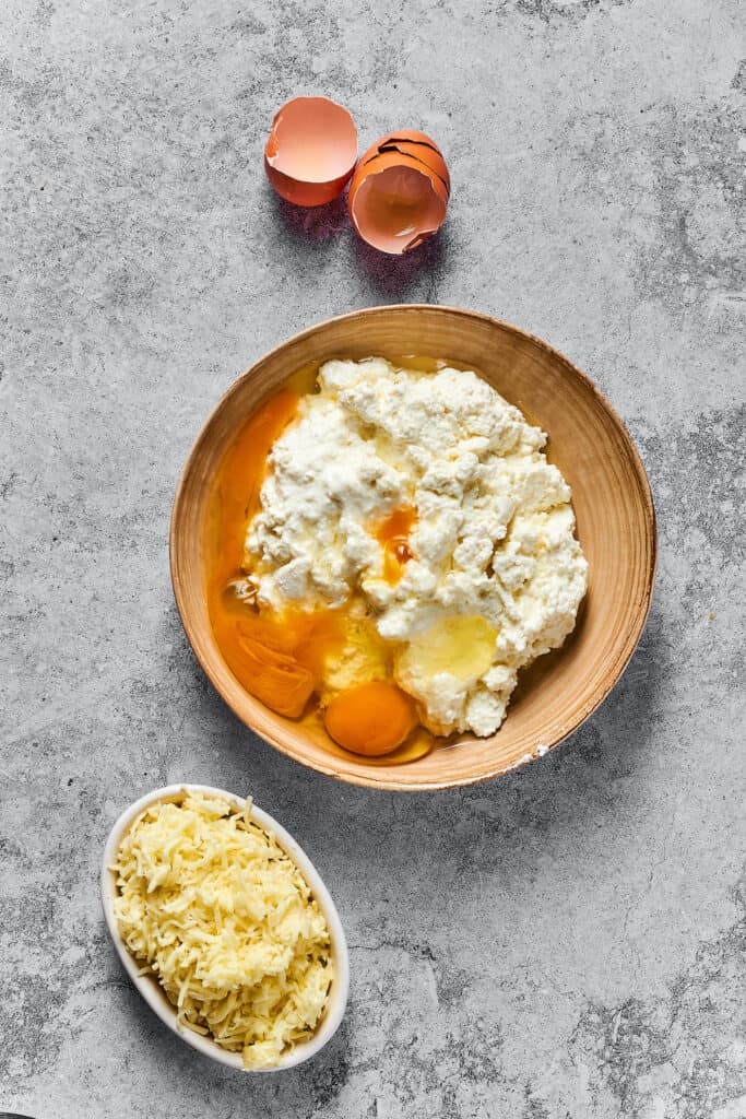 Cottage cheese, parmesan cheese, eggs, and other cheese filling ingredients in a bowl. A smaller bowl of shredded mozzarella is nearby on the work surface.