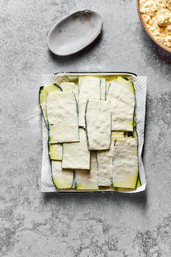Strips of raw zucchini, lightly-floured and resting on a paper-towel-lined tray.
