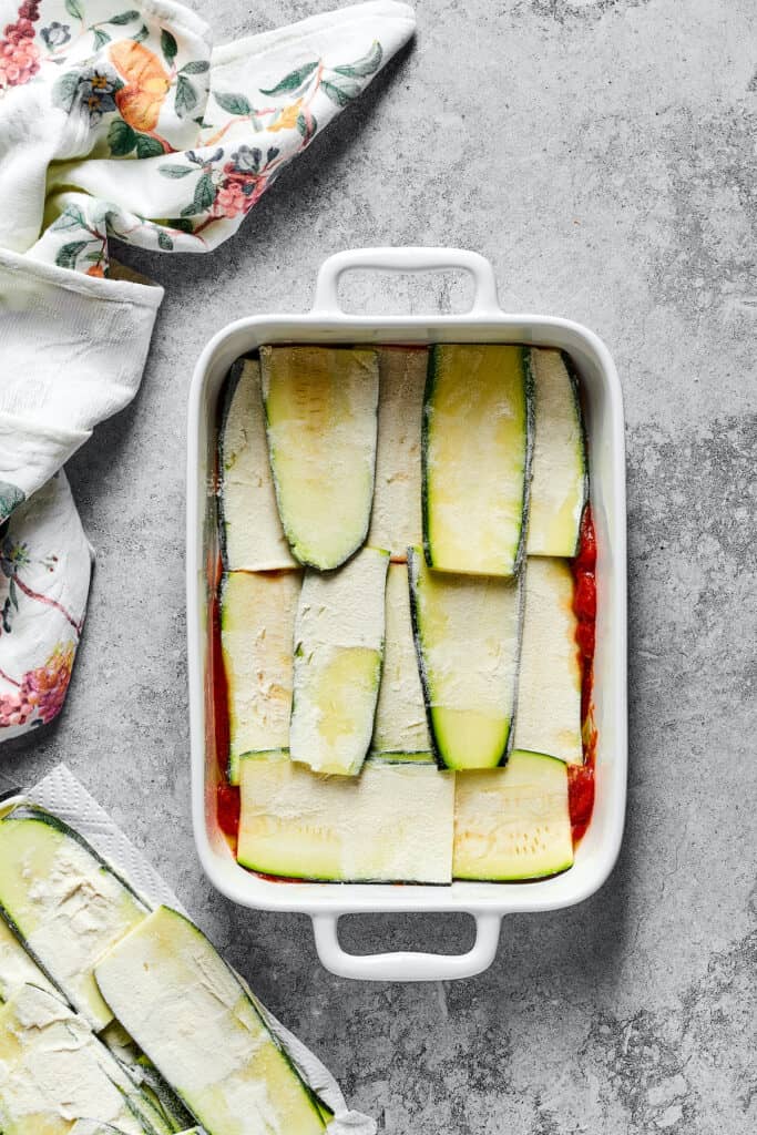 Strips of floured zucchini layered over a base of tomato sauce in a baking dish.