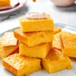 A platter of cornbread with some squares stacked on each other. The top piece of cornbread has a dollop of cinnamon cream on top and is being drizzled with honey.