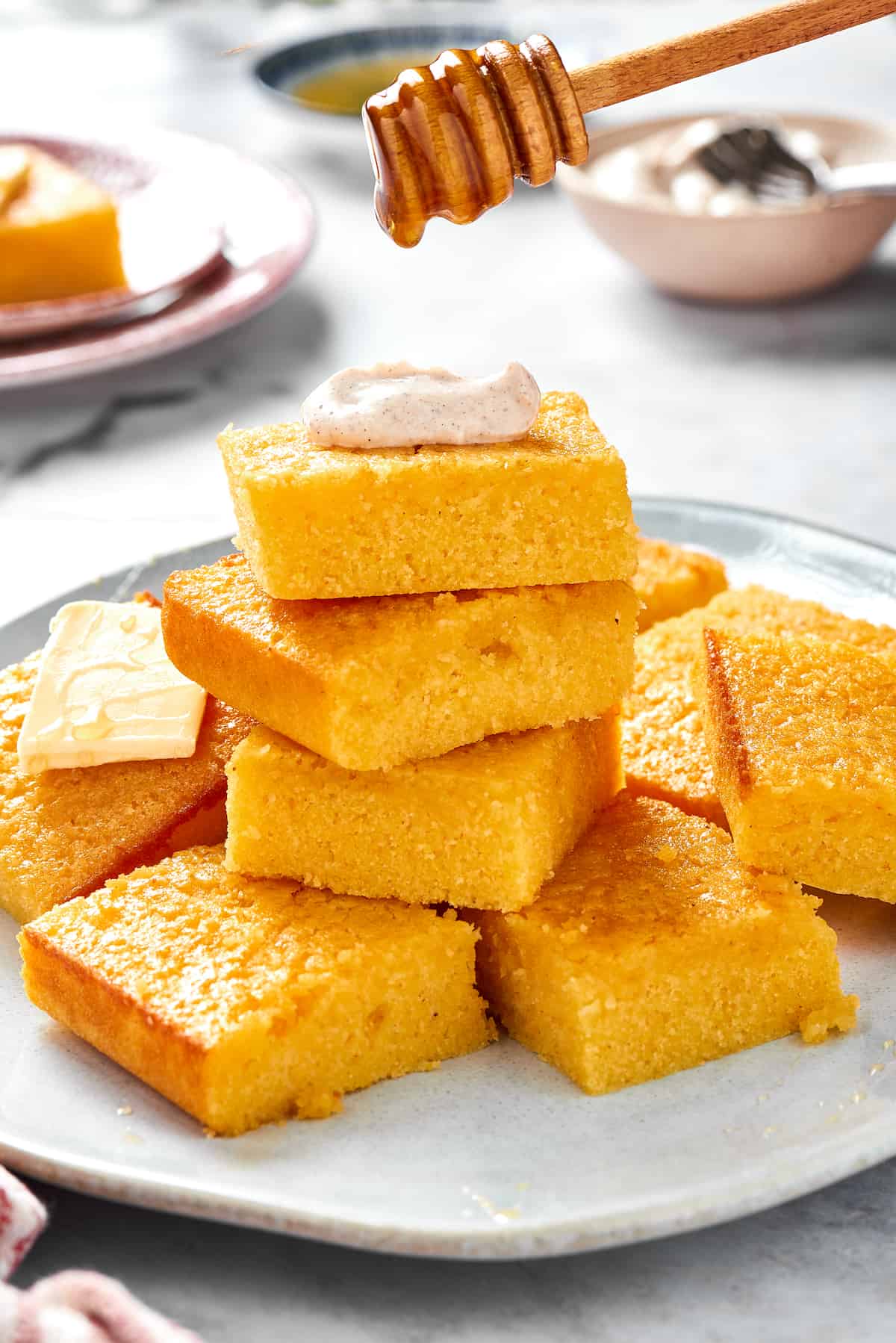 A platter of cornbread with some squares stacked on each other. The top piece of cornbread has a dollop of cinnamon cream on top and is being drizzled with honey.