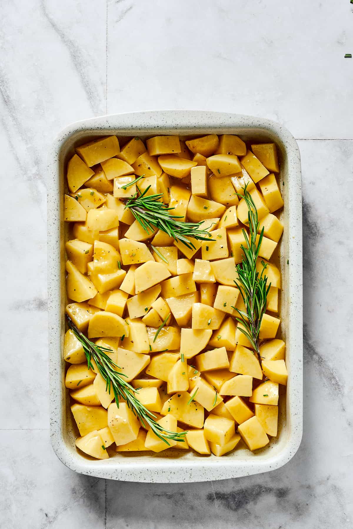 Seasoned raw potatoes and rosemary sprigs in a baking dish.