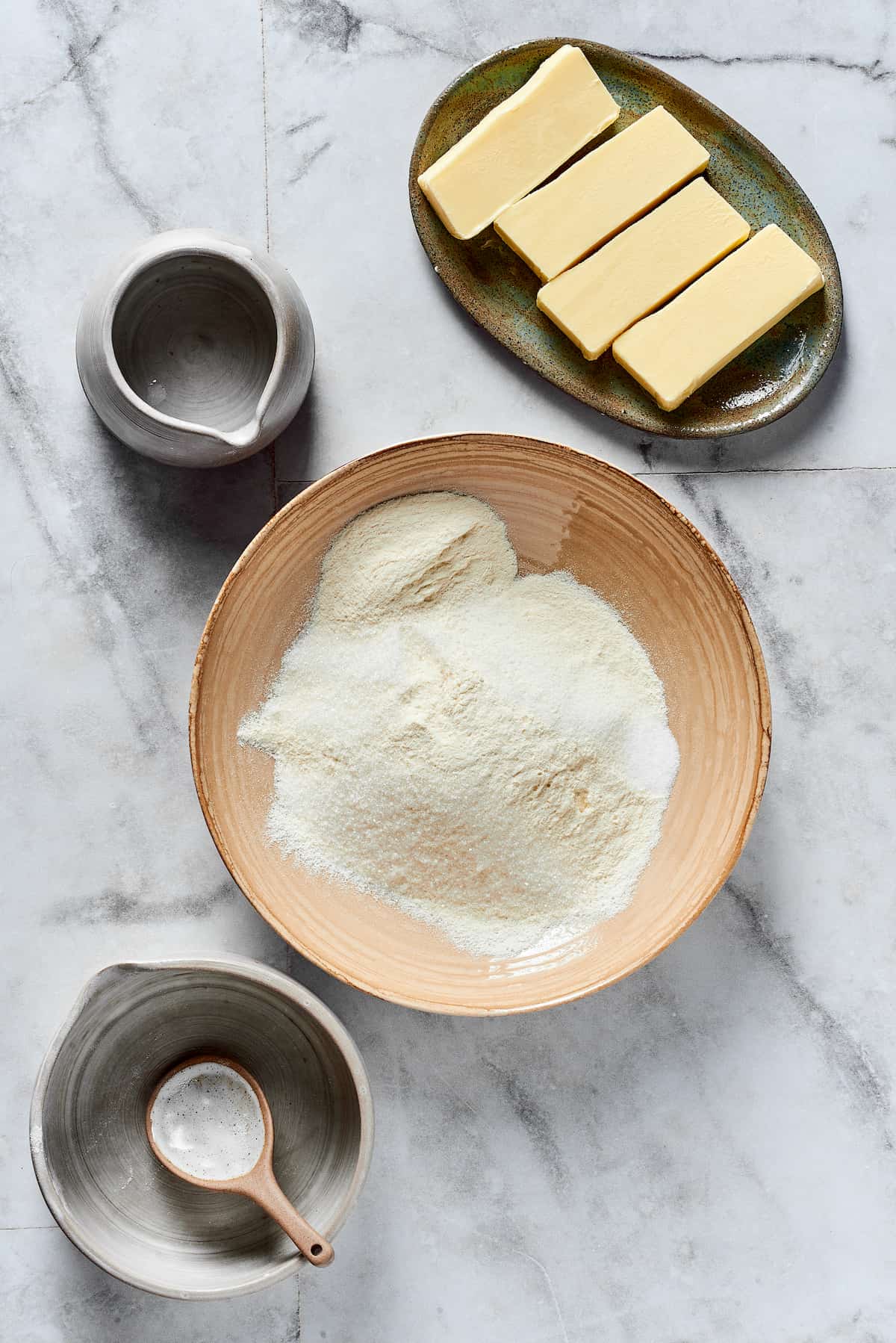 A mixing bowl with dry baking ingredients in it. Arranged around the mixing bowl are dishes of butter and ice water.