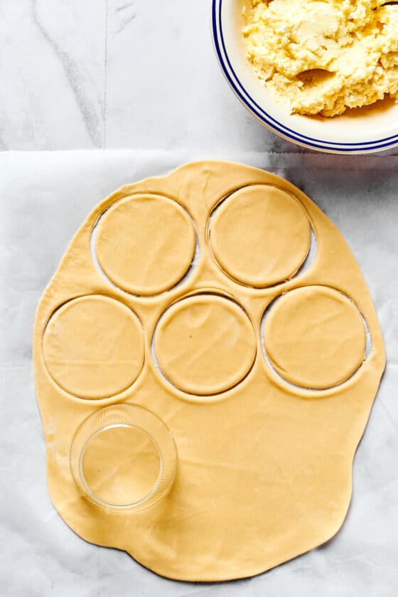 A sheet of rolled out dough on a work surface, with rounds cut out of it. A bowl of cheesy potato filling is nearby.