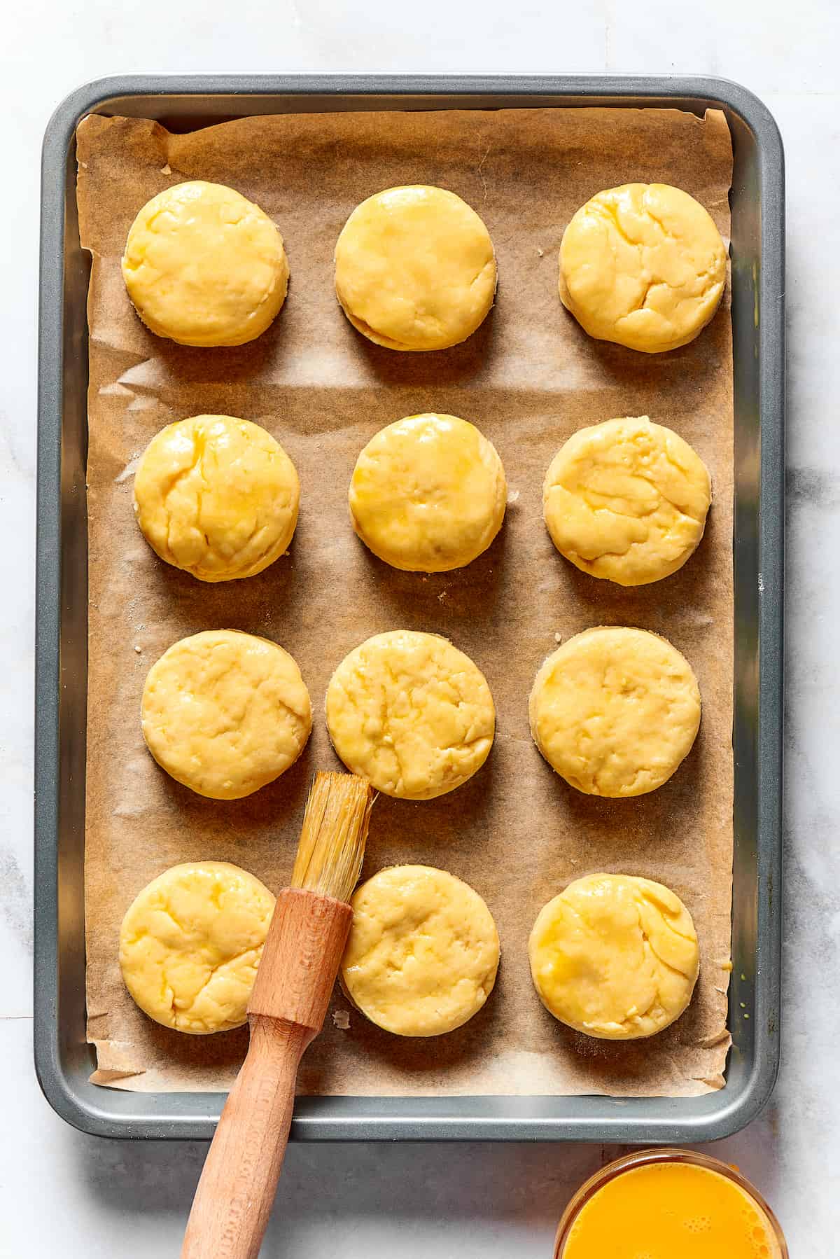 A dozen unbaked biscuits on a baking sheet, being brushed with beaten egg.