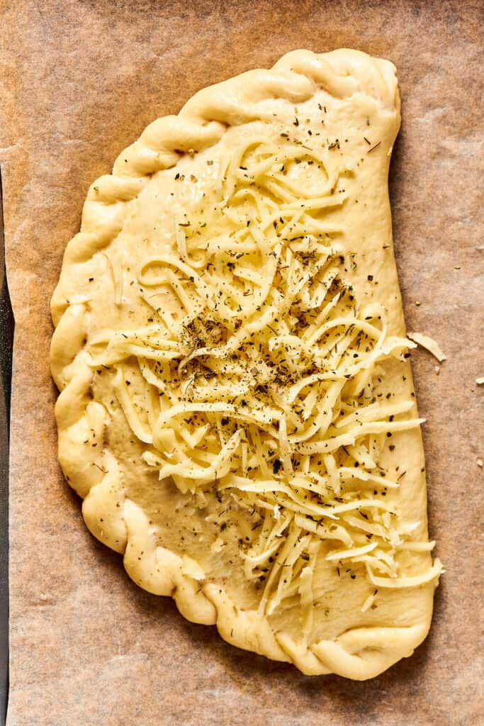 An unbaked calzone topped with shredded cheese and seasoning.