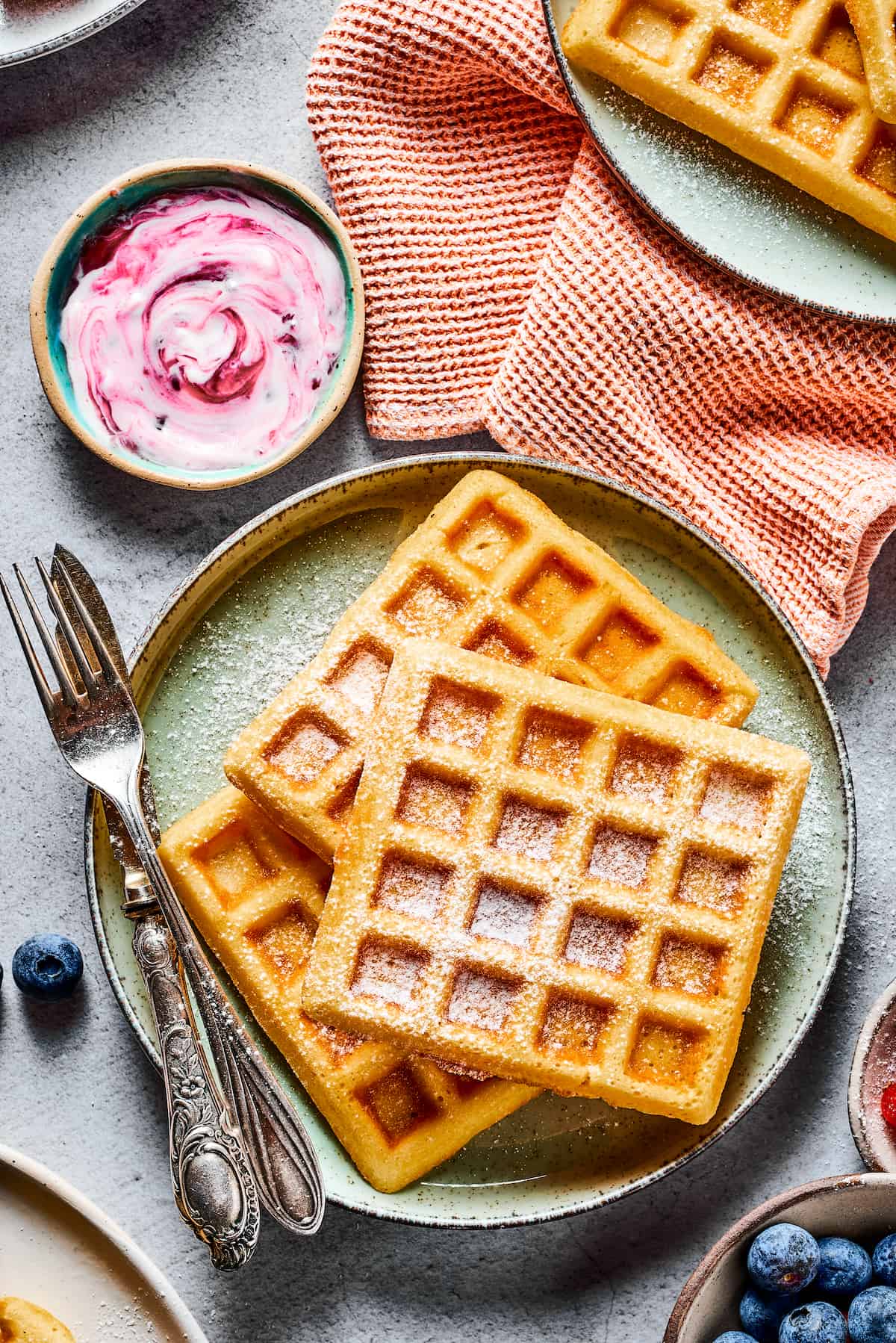 Overhead shot of three waffles on a plate, with a cloth napkin, cup of yogurt, and dish of fresh fruit.