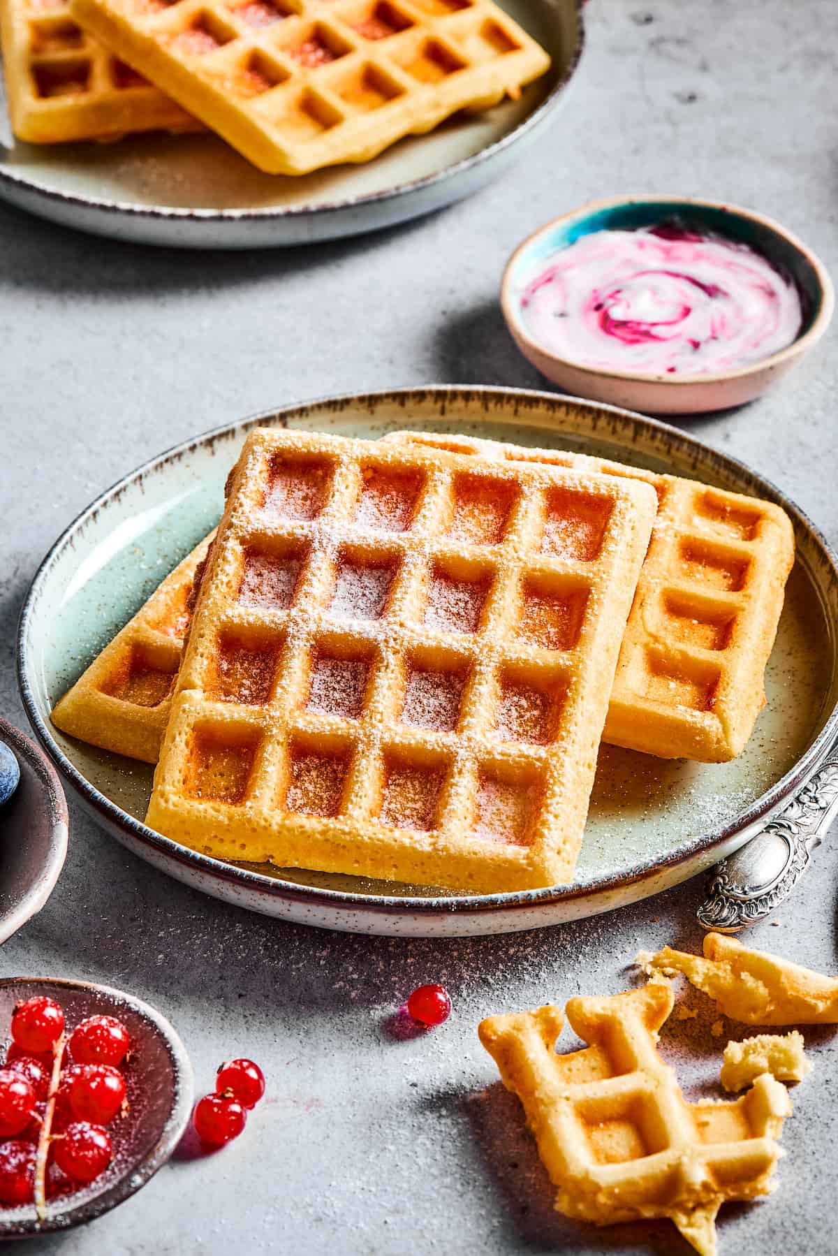 Plates of waffles on a gray work surface with dishes of berry yogurt and currants.