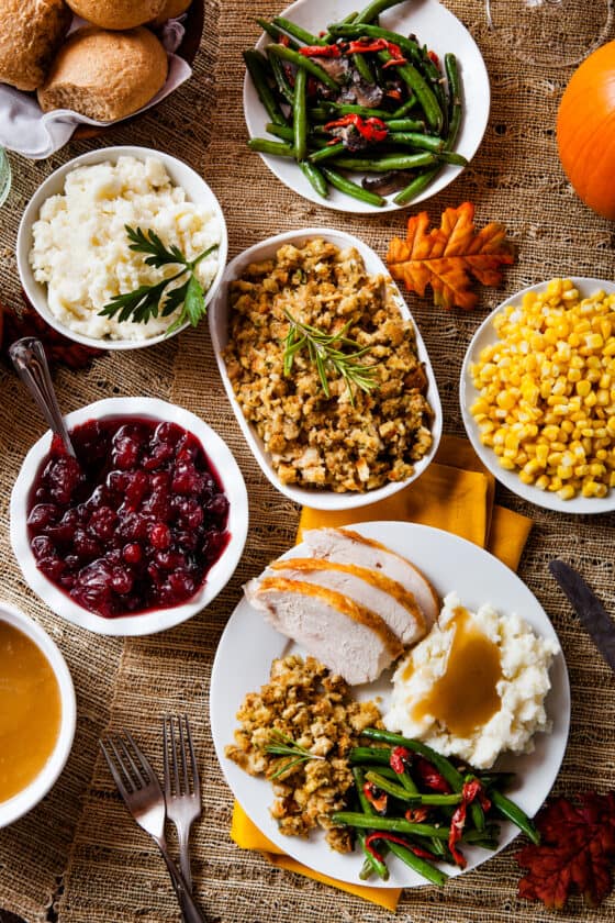 an overhead shot of a variety of serving plates filled with thanksgiving sides. From bottom left: gravy bowl, cranberry sauce in a white bowl, mashed potatoes, stuffing, green beans, corn, and a plate with sliced turkey, mashed potatoes, green beans, and corn.