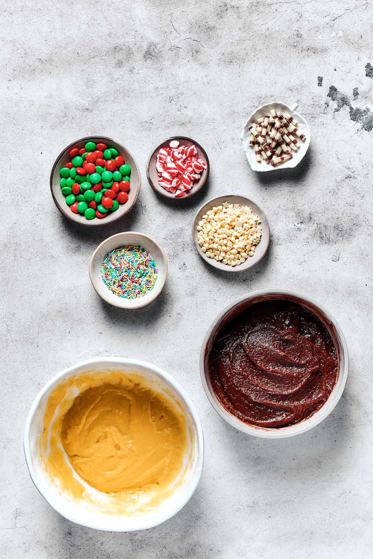 Two bowls of cookie dough next to small dishes of decorations. one of the bowls is chocolate, one is vanilla.