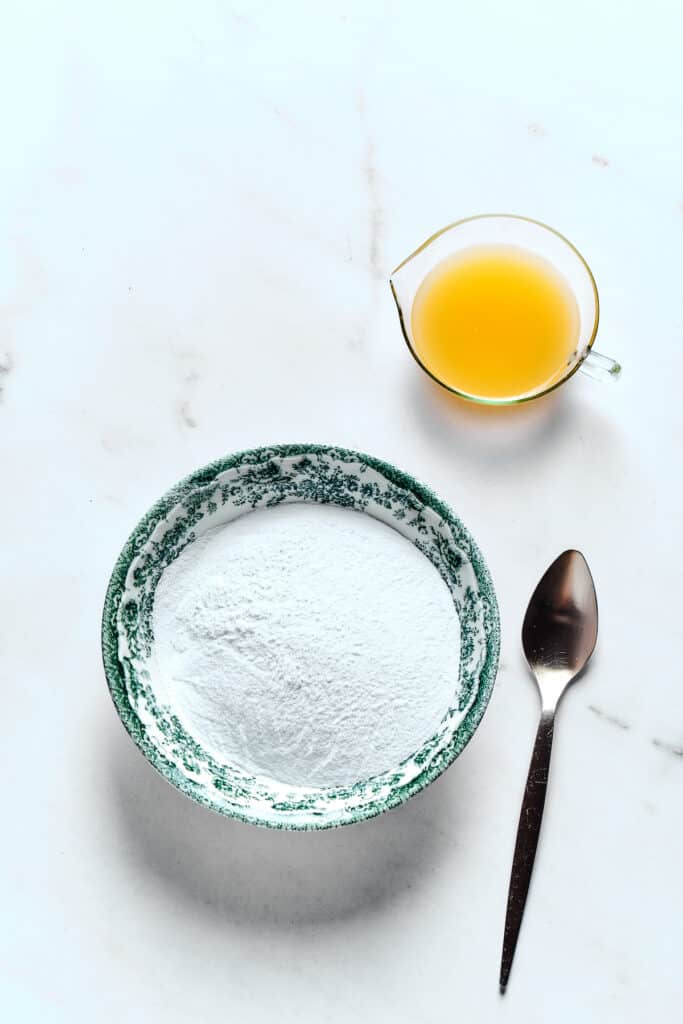 A bowl of powdered sugar next to a glass of orange juice and a spoon.