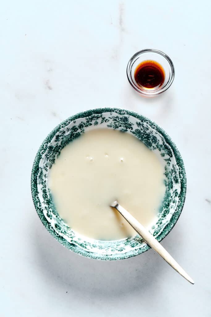 A bowl of creamy glaze with a small dish of vanilla. A spoon is resting in the bowl.