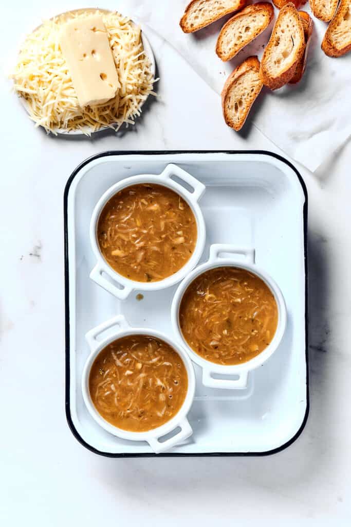 Individual oven-proof bowls of French onion soup next to baguette slices and a dish of shredded cheese.