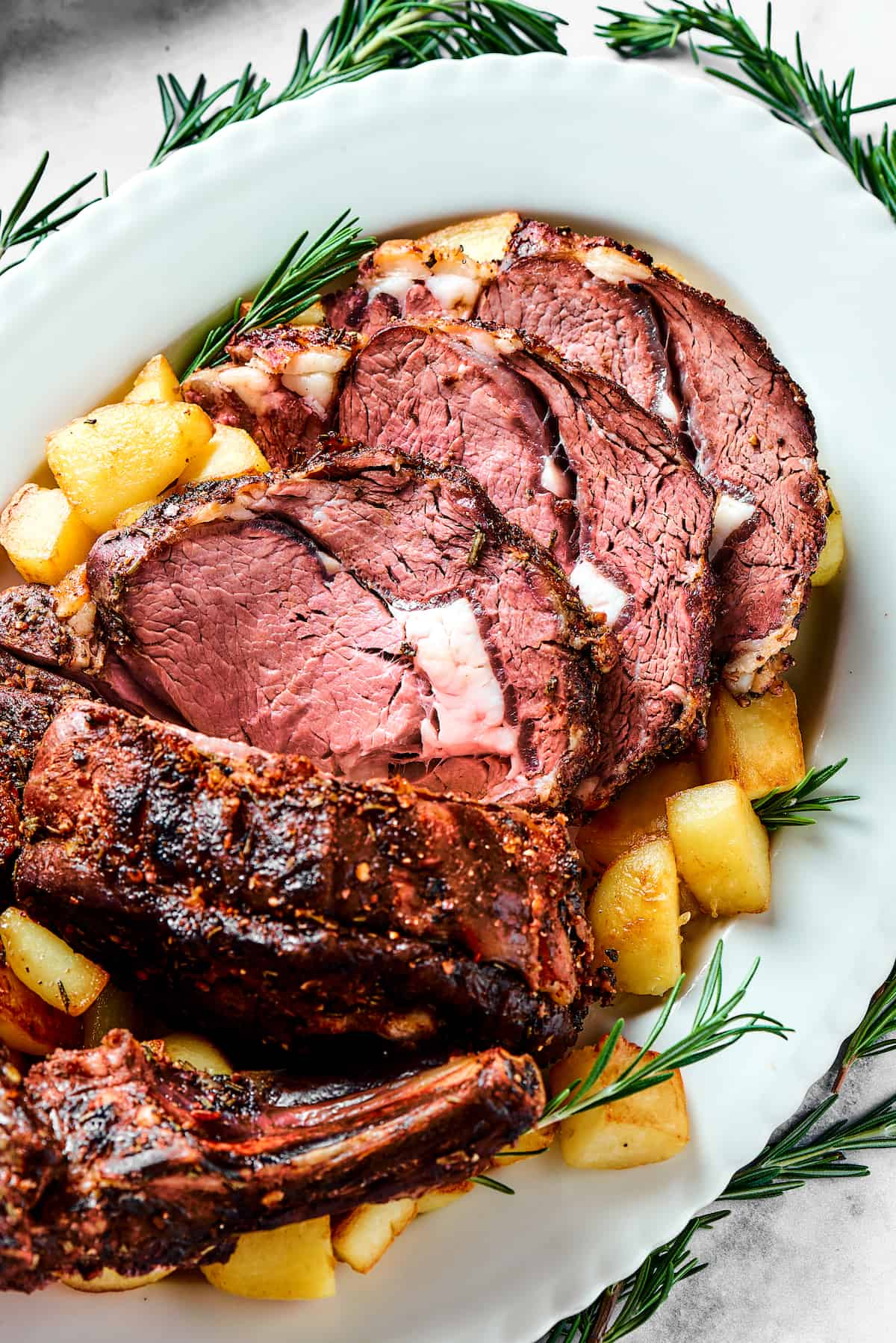 Sliced standing rib roast on a serving patter with potatoes and herbs.