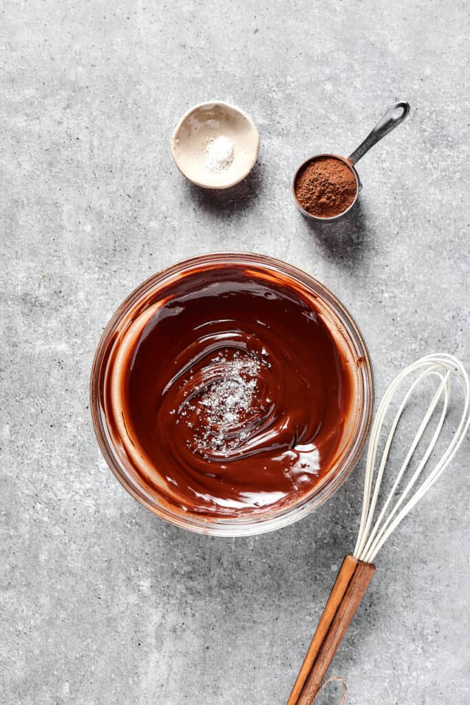 A bowl of melted butter and chocolate next to a measuring cup of cocoa powder and a dish of salt.