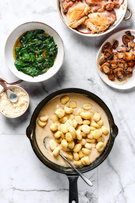 Gnocchi and cream sauce in a skillet, next to dishes of spinach, mushrooms, and chicken.