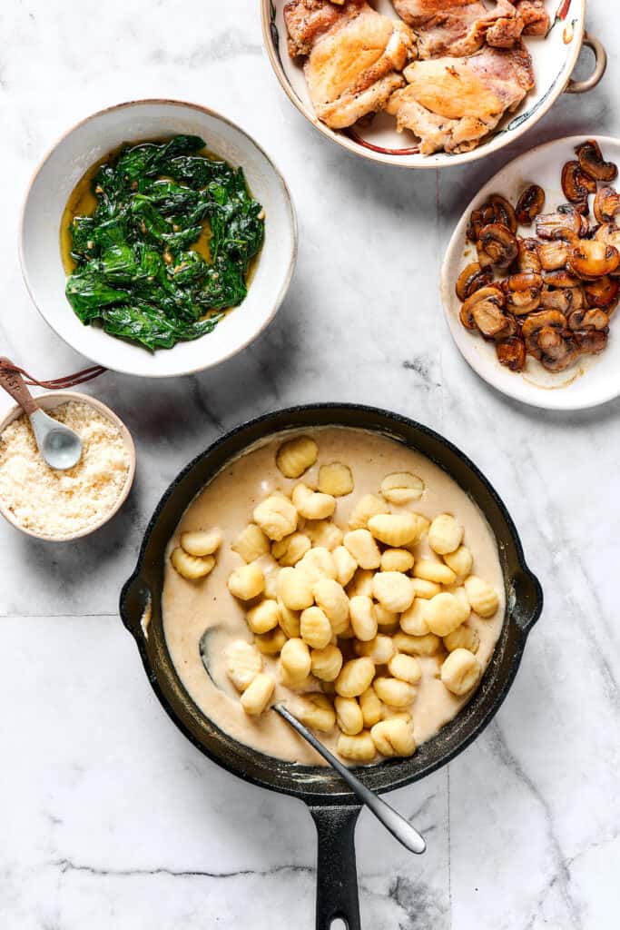 Gnocchi and cream sauce in a skillet, next to dishes of spinach, mushrooms, and chicken.