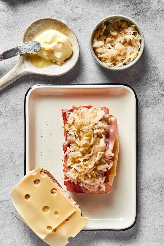 One slice of bread topped with cheese, corned beef, and sauerkraut, next to another slice with just cheese.