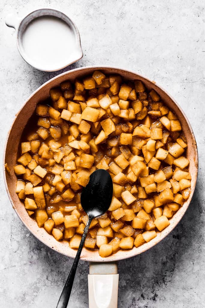 cooking diced apples in a skillet, with a dark spoon set inside the skillet, and a cup of milk placed next to the skillet.