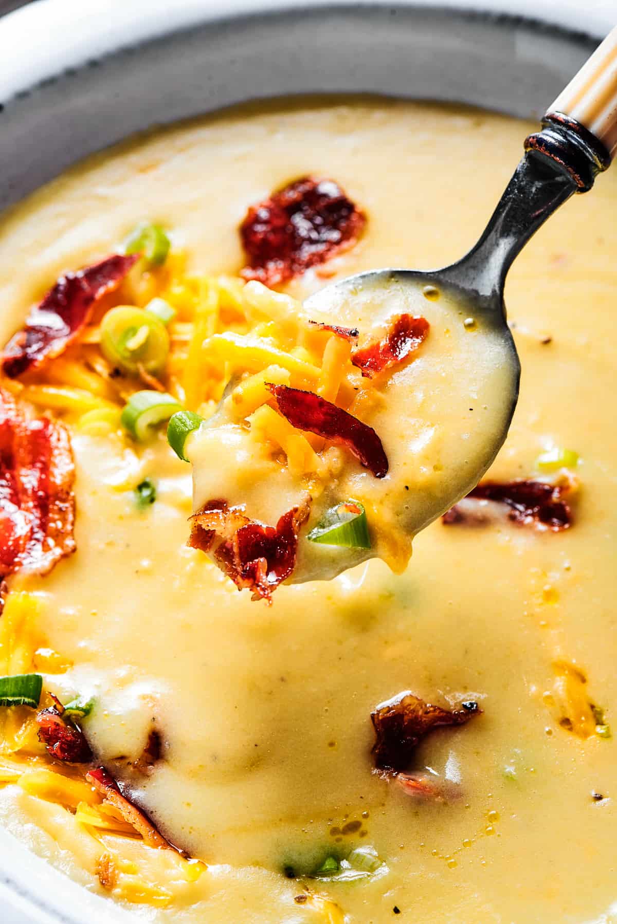 A white bowl of loaded potato soup, with a spoon lifted out of the bowl toward the camera.