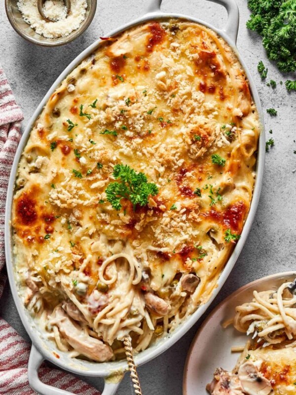 A large chicken tetrazzini casserole next to a dinner plate and a bunch of fresh herbs.
