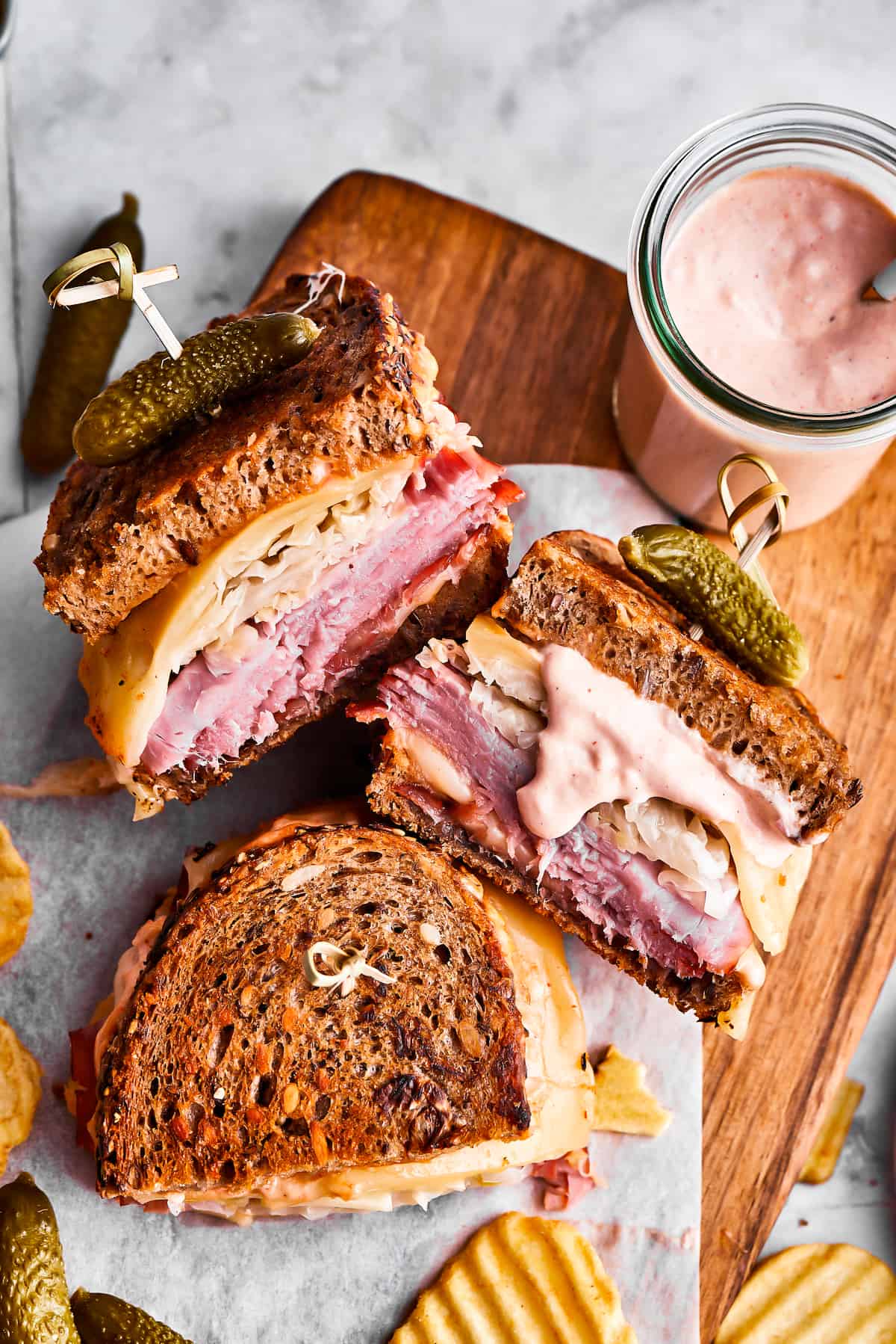 Three Reuben sandwich halves, some potato chips, and a jar of dressing on a cutting board.