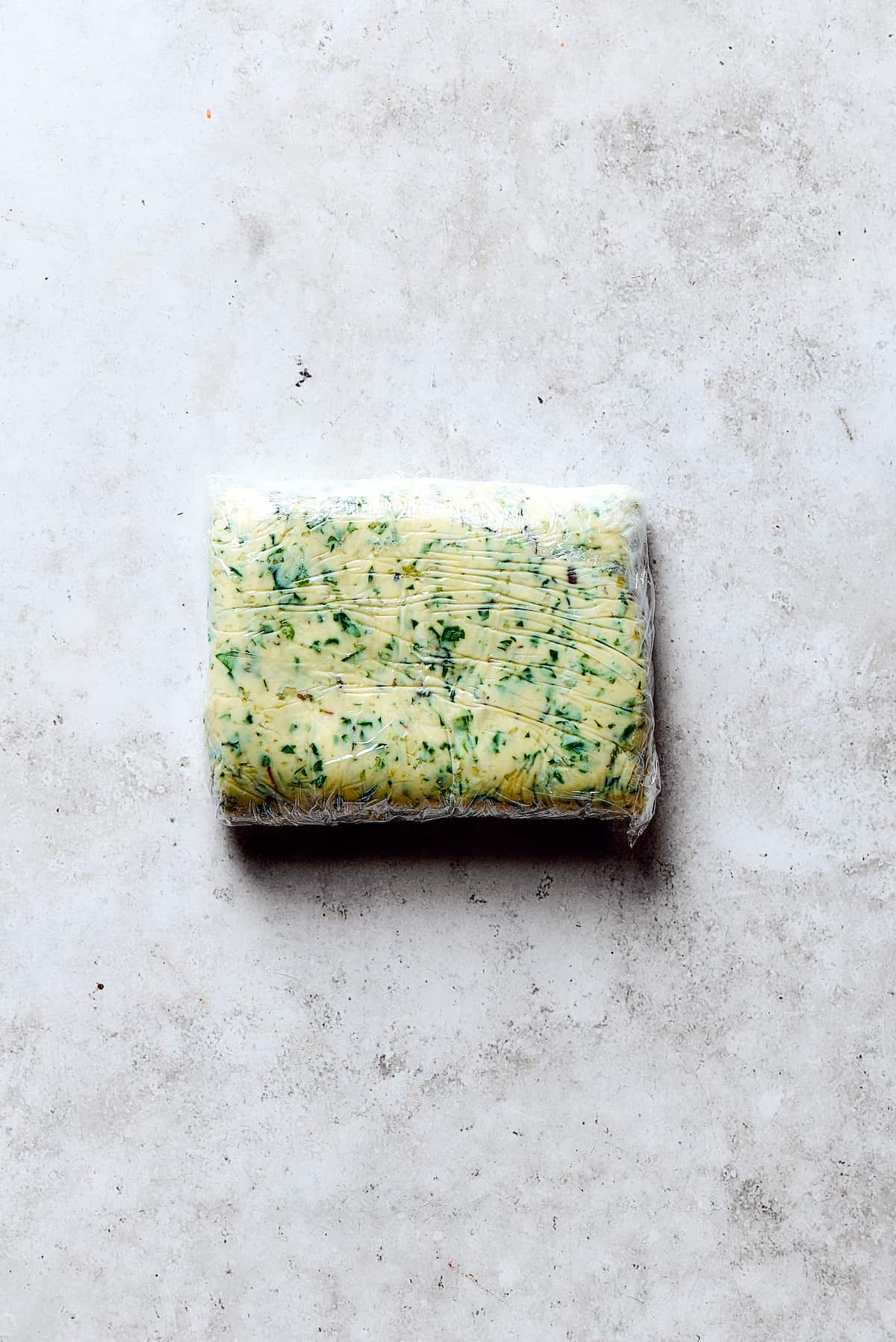 A block of frozen garlic herb butter, wrapped in plastic wrap, indicating it's ready for slicing and stuffing.