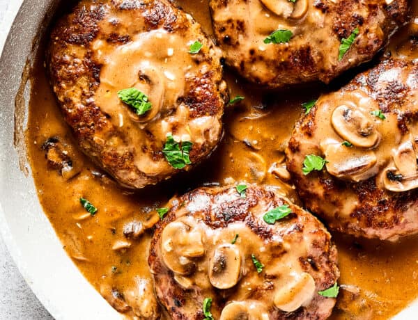 Four salisbury steaks in gravy, in a skillet. Vegetables, herbs, and a cloth napkin are arranged around the skillet.