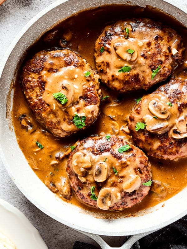 Four salisbury steaks in gravy, in a skillet. Vegetables, herbs, and a cloth napkin are arranged around the skillet.