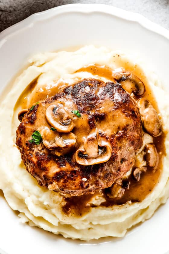 Salisbury steak with mushroom gravy served on a dinner plate over mashed potatoes.