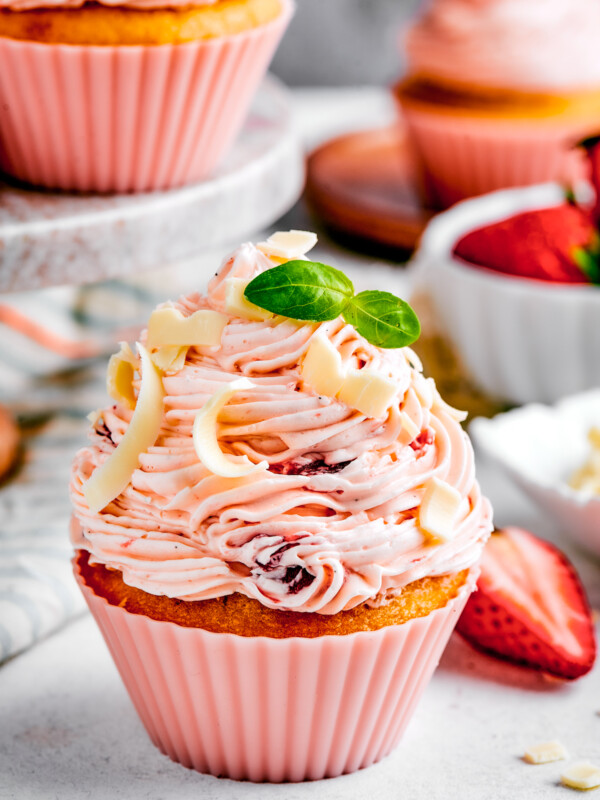 A strawberry cupcake in a pink cupcake liner with a tiny sprig of mint on top. More cupcakes are visible in the background of the shot.