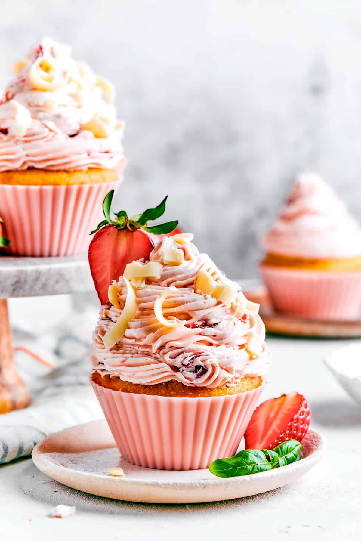 A strawberry cupcake in a pink cupcake liner with a tiny sprig of mint on top. More cupcakes are visible in the background of the shot.