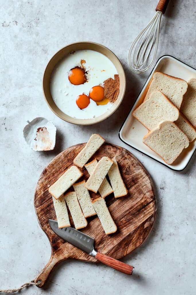 A bowl of eggs, milk, and other custard ingredients next to a tray of sliced bread, and a cutting board of additional bread cut into sticks.