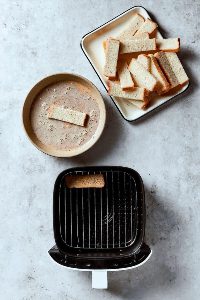 An overhead shot of an air fryer, a bowl of custard, and sticks of bread. One piece of bread is in the custard. A custard-soaked piece is in the air fryer, as well.