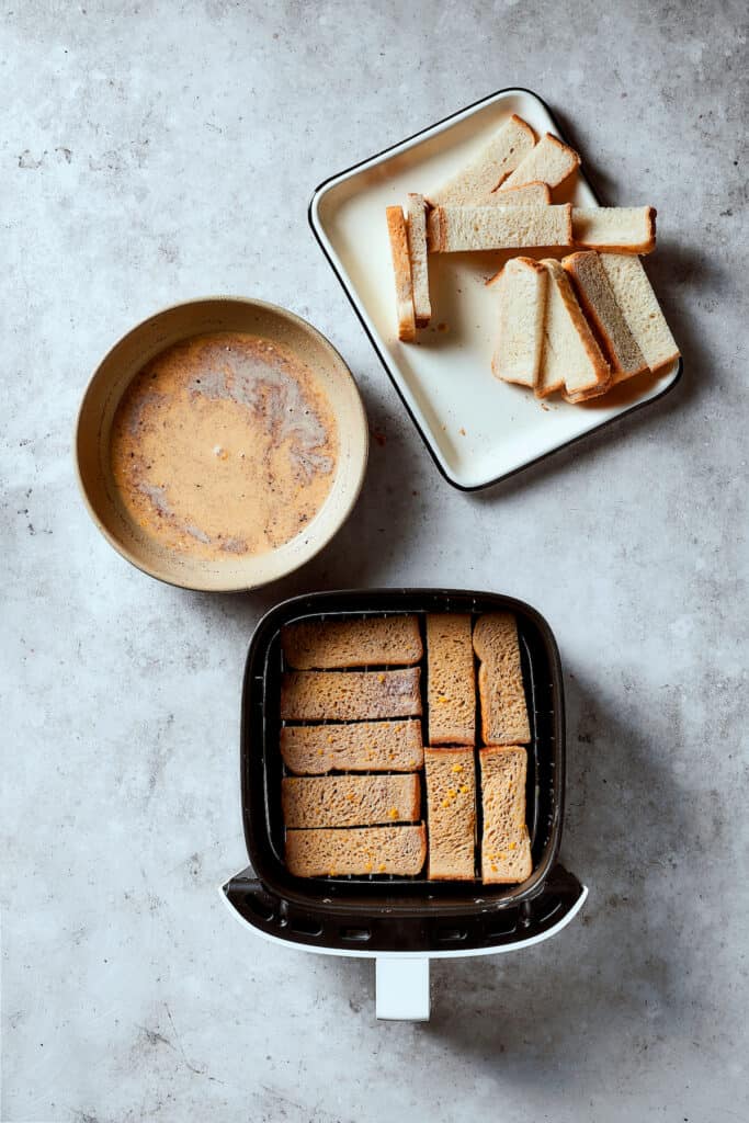 Custard-soaked bread sticks lined up neatly in an air fryer. Additional bread and custard are nearby.