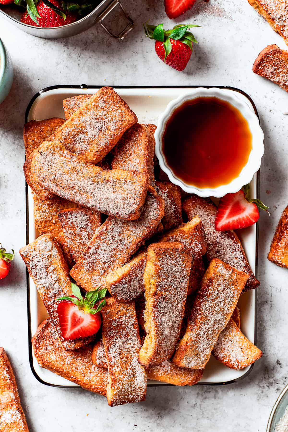 Air fryer French toast sticks on an enamel tray with a cup of maple syrup and fresh strawberries.