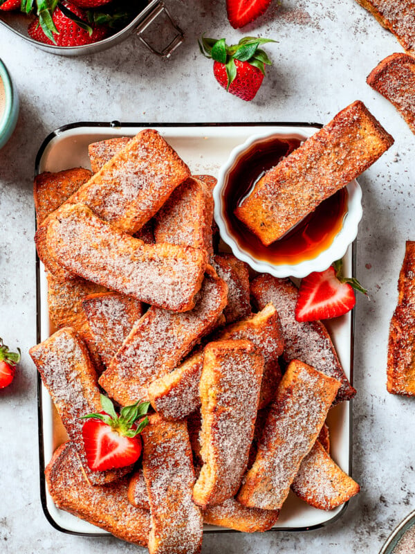 A tray of French toast sticks with maple syrup and strawberries. One stick is dunked in the syrup.