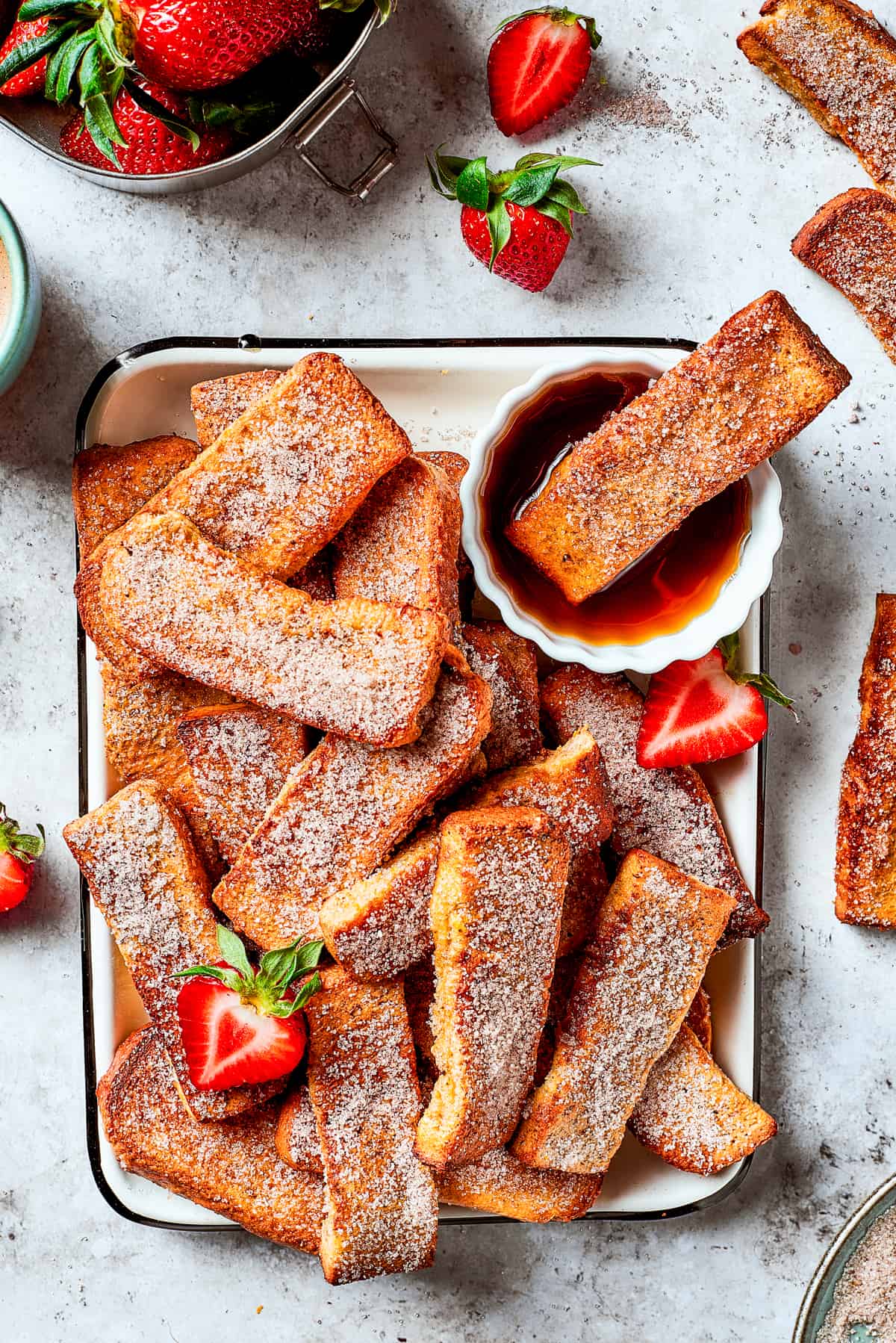 A tray of French toast sticks with maple syrup and strawberries. One stick is dunked in the syrup.