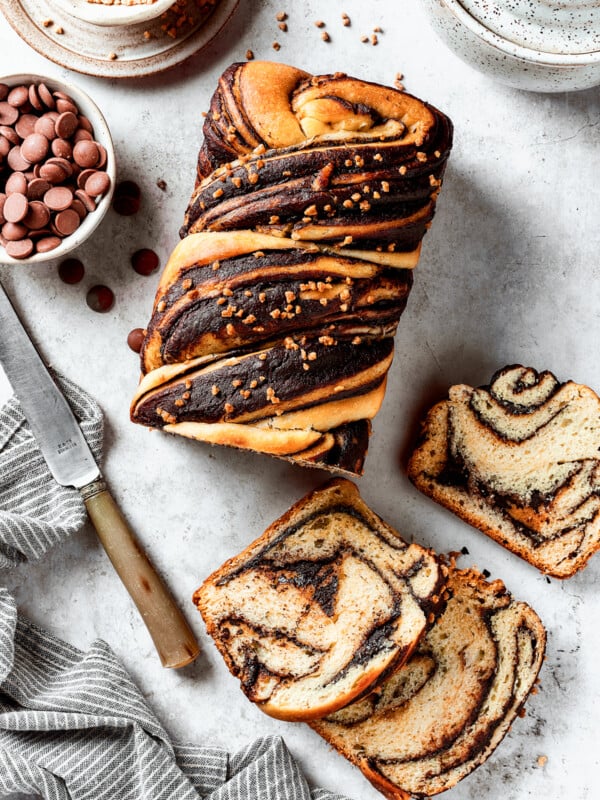 A loaf of chocolate babka that has been partially sliced. The slices are laid out on the work surface along with a bread knife and a dish of chocolate chips.
