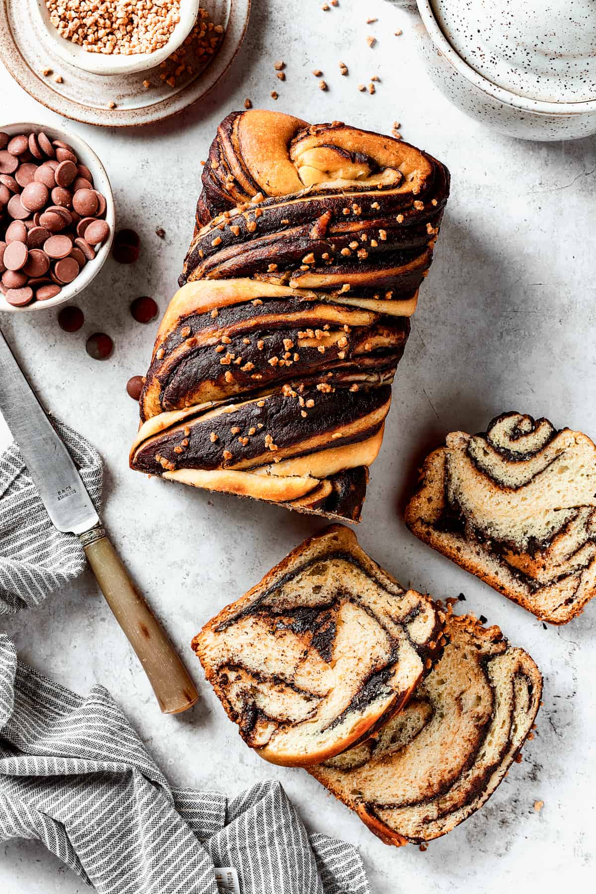 A loaf of chocolate babka that has been partially sliced. The slices are laid out on the work surface along with a bread knife and a dish of chocolate chips.
