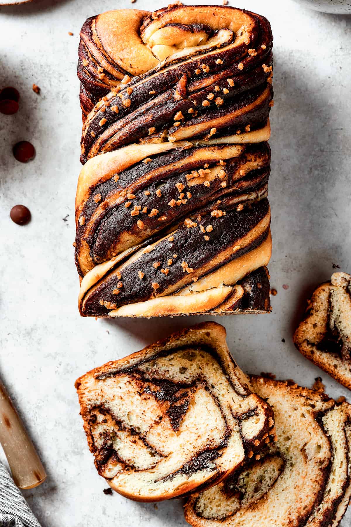 A loaf of chocolate babka that has been partially sliced. The slices are laid out on the work surface, along with a bread knife and a dish of chocolate chips.