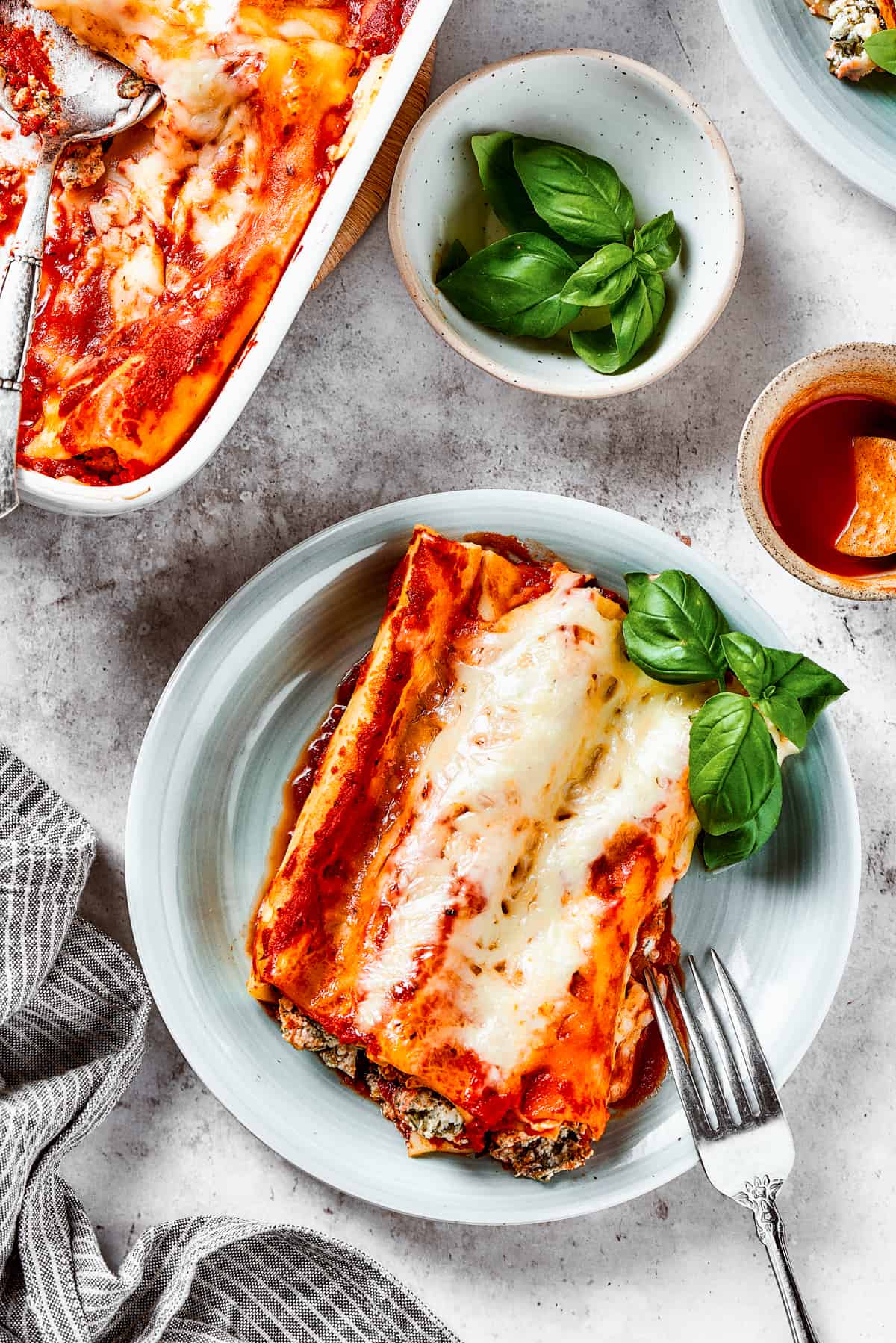 Overhead shot of stuffed manicotti on a plate, next to a casserole dish with a serving spoon resting in it.